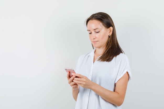 Junge Dame, die ihr Telefon in der weißen Bluse betrachtet und konzentriert schaut