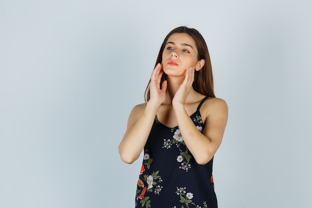 Junge Dame, die ihre Gesichtshaut mit den Händen in der Bluse untersucht und nachdenklich aussieht, Vorderansicht.