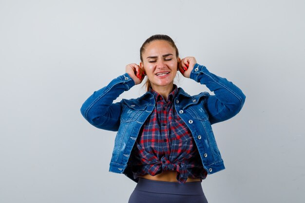 Junge Dame, die ihre Ohrläppchen in kariertem Hemd, Jeansjacke herunterzieht und lustig aussieht. Vorderansicht.