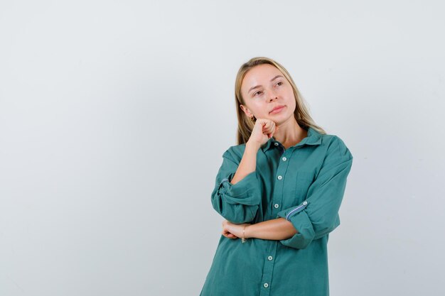 Junge Dame, die im grünen Hemd das Kinn auf die Faust stützt und nachdenklich aussieht looking