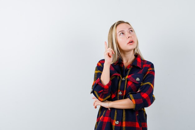 Junge Dame, die im karierten Hemd nach oben zeigt und nachdenklich aussieht. Vorderansicht.