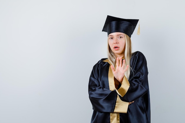 Junge Dame, die in akademischer Kleidung eine Stopp-Geste zeigt und verängstigt aussieht