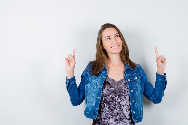 Junge Dame, die in Bluse, Jeansjacke nach oben zeigt und glücklich aussieht, Vorderansicht.