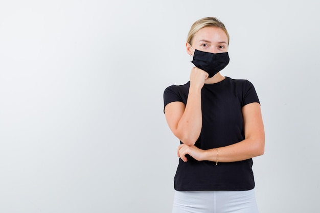 Junge Dame, die in denkender Pose in T-Shirt, Hose, medizinischer Maske steht und selbstbewusst aussieht