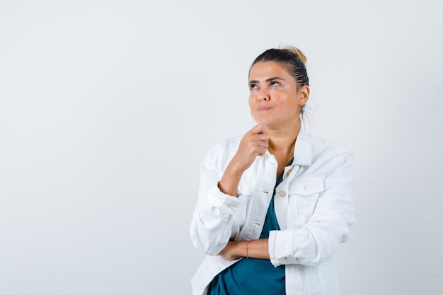 Junge Dame, die in der denkenden Haltung in der weißen Jacke steht und verträumte Vorderansicht schaut.