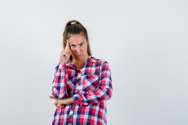 Junge Dame, die in der denkenden Pose im karierten Hemd steht und ernsthafte Vorderansicht schaut.