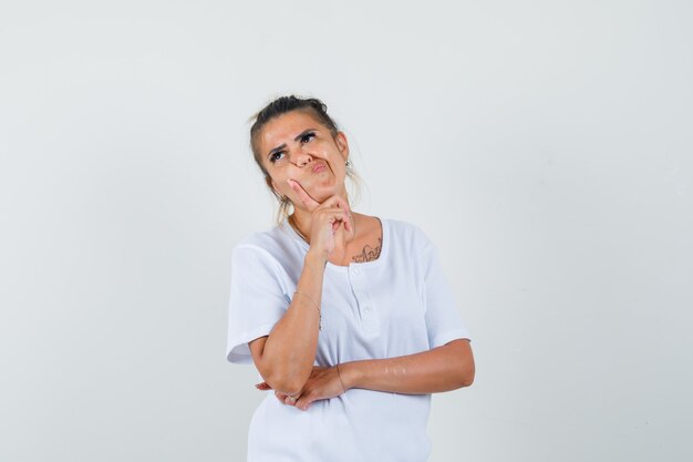 Junge Dame, die in der denkenden Pose im T-Shirt steht und zögernd schaut