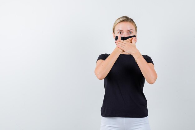 Junge Dame, die in T-Shirt, Hose, medizinischer Maske die Hände am Mund hält und verwirrt aussieht