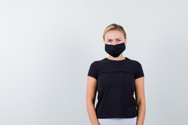 Junge Dame, die in T-Shirt, Hose, medizinischer Maske in die Kamera schaut und wehmütig aussieht