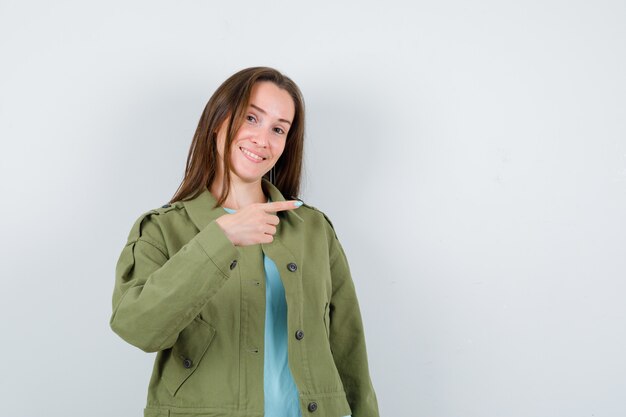 Junge Dame, die in T-Shirt, Jacke auf die rechte Seite zeigt und fröhlich aussieht. Vorderansicht.