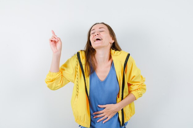 Junge Dame, die in T-Shirt, Jacke nach oben zeigt und glückselig aussieht, Vorderansicht.