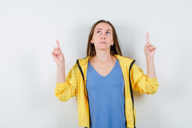 Junge Dame, die in T-Shirt, Jacke nach oben zeigt und hoffnungsvoll aussieht, Vorderansicht.