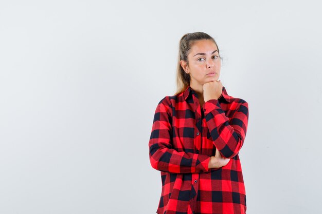 Junge Dame, die Kinn auf Faust im karierten Hemd stützt und nachdenklich aussieht