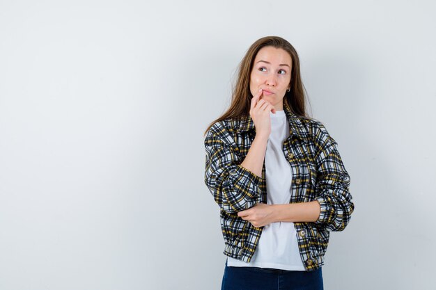 Junge Dame, die Kinn auf Hand in T-Shirt, Jacke stützt und zögernd, Vorderansicht schaut.