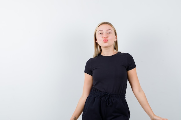 Junge Dame, die Kuss mit schmollenden Lippen im T-Shirt, in der Hose und im niedlichen Blick sendet. Vorderansicht.