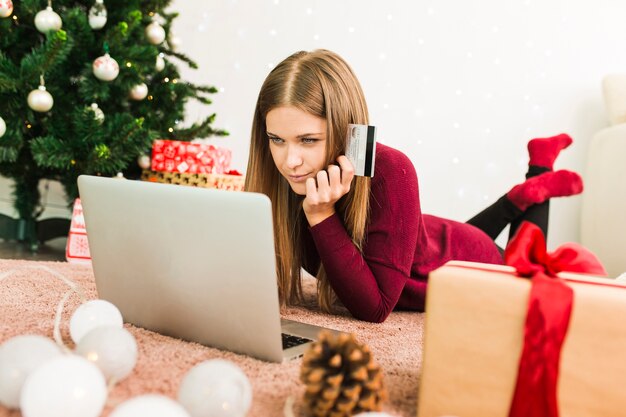 Junge Dame, die Laptop mit Kreditkarte nahe Geschenkboxen und Weihnachtstannenbaum verwendet