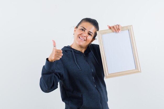 Junge Dame, die leeren Rahmen hält, während Daumen oben in Jacke zeigt und optimistisch schaut. Vorderansicht.