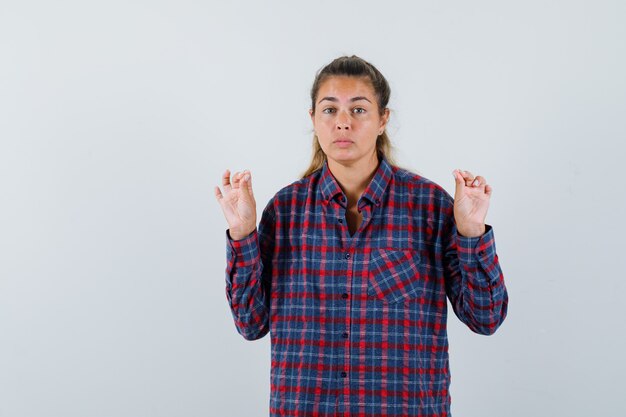 Junge Dame, die Meditationsgeste im karierten Hemd zeigt und hoffnungsvoll aussieht. Vorderansicht.