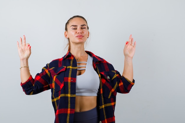 Junge Dame, die Meditationsgeste in der Spitze zeigt, kariertes Hemd und entspannt aussieht, Vorderansicht.