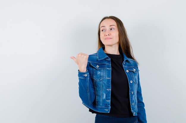 Junge Dame, die mit dem Daumen in Bluse, Jacke zurück zeigt und selbstbewusst aussieht, Vorderansicht.