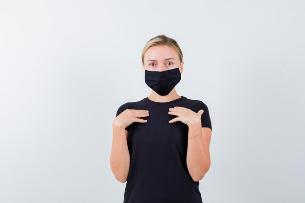 Junge Dame, die mit den Händen in T-Shirt, Hose, medizinischer Maske auf sich selbst zeigt