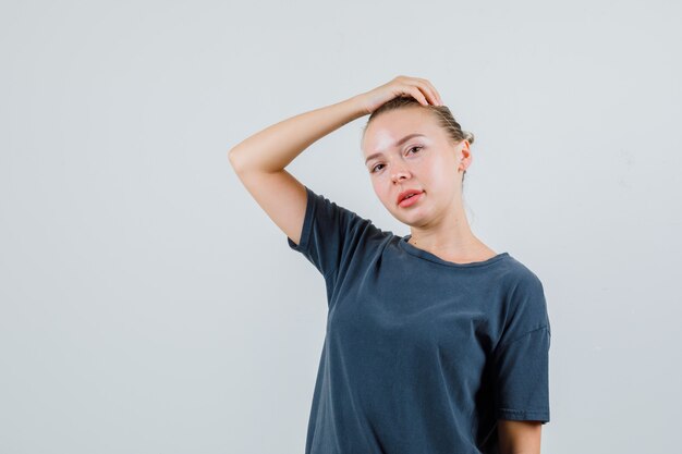 Junge Dame, die mit erhabener Hand auf Kopf im grauen T-Shirt aufwirft und niedlich schaut