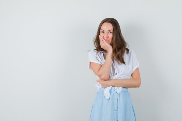 Junge Dame, die mit Gesicht lehnender Hand in der weißen Bluse, im blauen Rock steht und zögernd schaut