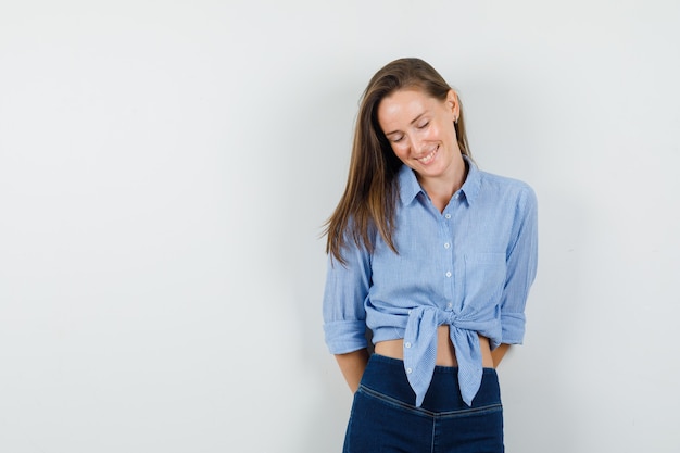 Junge Dame, die mit Händen auf ihrem Rücken im blauen Hemd, in der Hose unten schaut und optimistisch schaut