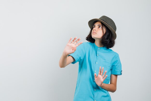 Junge Dame, die mit Händen im blauen Hemd, Hut verteidigt