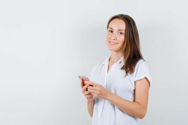 Junge Dame, die mit Telefon in der weißen Bluse aufwirft und schön schaut