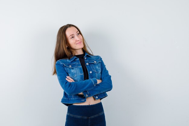 Junge Dame, die mit verschränkten Armen in Bluse, Jacke, Jeans steht und verträumt aussieht.