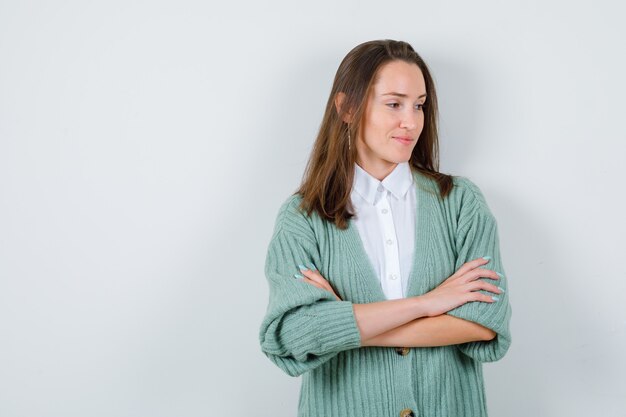 Junge Dame, die mit verschränkten Armen in Hemd, Strickjacke steht und zufrieden aussieht. Vorderansicht.