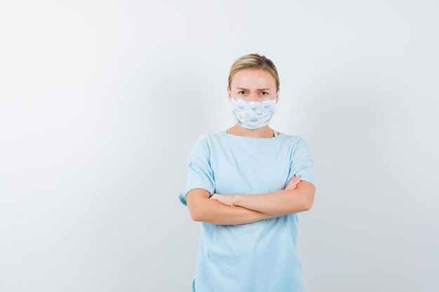 Junge Dame, die mit verschränkten Armen in T-Shirt, Maske steht und verärgert aussieht