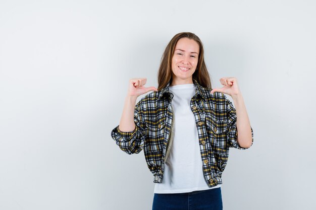 Junge Dame, die mittlere Daumen im T-Shirt, in der Jacke, in den Jeans zeigt und glücklich schaut, Vorderansicht.