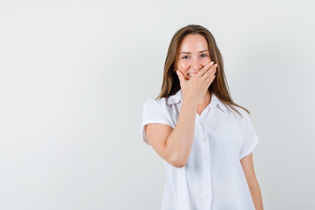 Junge Dame, die Mund mit Hand bedeckt, während sie in der weißen Bluse lacht und fröhlich aussieht