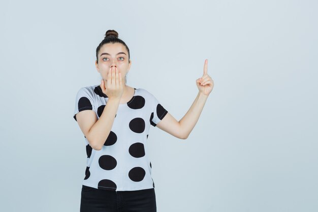 Junge Dame, die nach oben zeigt, während Mund mit Hand in T-Shirt, Jeans bedeckt und verwirrt aussieht. Vorderansicht.