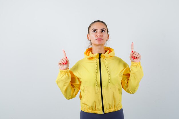 Junge Dame, die nach oben zeigt, während sie im Trainingsanzug nach oben schaut und hoffnungsvoll aussieht, Vorderansicht.