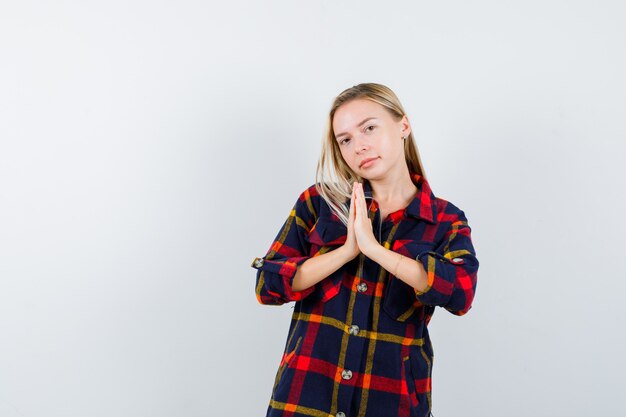 Junge Dame, die namaste Geste im karierten Hemd zeigt und hoffnungsvoll aussieht. Vorderansicht.