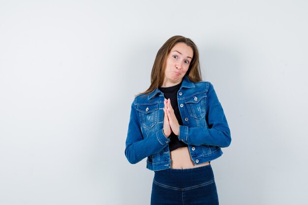 Junge Dame, die Namaste-Geste in Bluse, Jacke, Jeans zeigt und zögerlich aussieht, Vorderansicht.