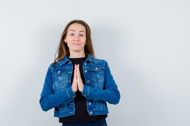 Junge Dame, die Namaste-Geste in Bluse, Jacke zeigt und hoffnungsvoll aussieht. Vorderansicht.