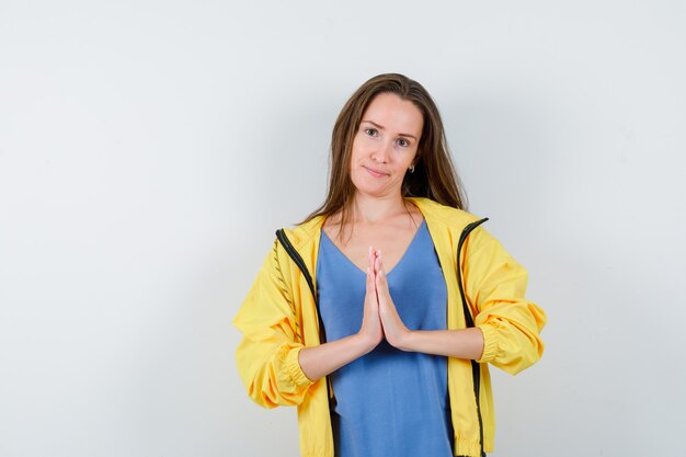 Junge Dame, die Namaste-Geste in T-Shirt, Jacke zeigt und hoffnungsvoll aussieht, Vorderansicht.