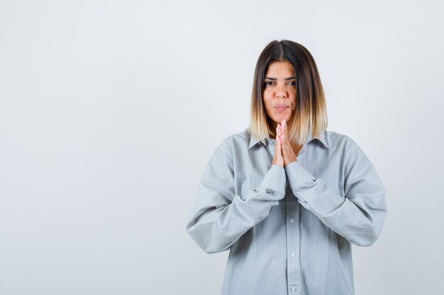 Junge Dame, die Namaste-Geste in übergroßem Hemd zeigt und friedlich aussieht. Vorderansicht.