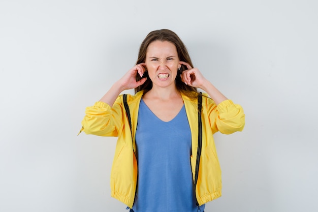 Junge Dame, die Ohren mit den Fingern im T-Shirt verstopft und gereizt schaut, Vorderansicht