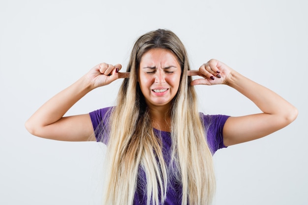 Junge Dame, die Ohren mit den Fingern im violetten T-Shirt verstopft und genervte Vorderansicht schaut.