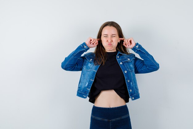 Junge Dame, die Ohren mit den Fingern in der Bluse verstopft und genervt aussieht, Vorderansicht.