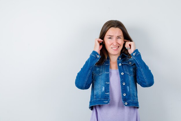 Junge Dame, die Ohren mit den Fingern in T-Shirt, Jacke verstopft und gelangweilt aussieht. Vorderansicht.