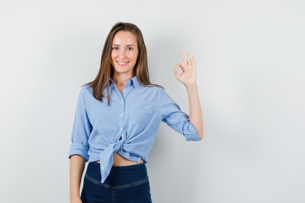 Junge Dame, die ok Geste im blauen Hemd, in der Hose und im fröhlichen Blick zeigt