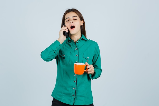 Junge Dame, die orange Tasse Tee hält, auf dem Handy im Hemd spricht und schläfrig sieht, Vorderansicht.