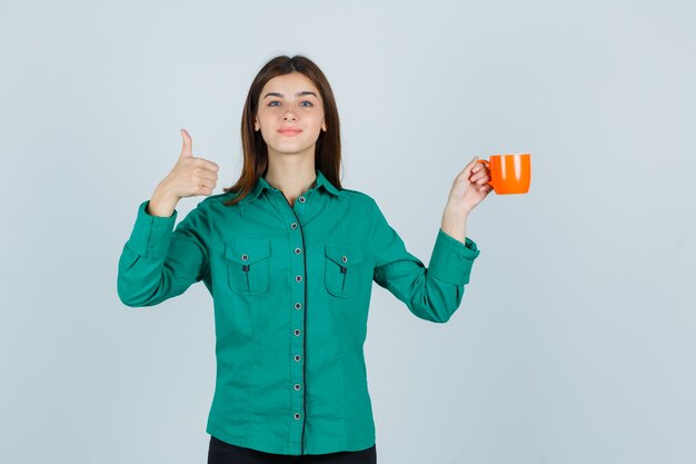 Junge Dame, die orange Tasse Tee hält, während Daumen oben im Hemd zeigt und erfreut aussieht. Vorderansicht.