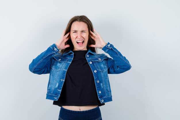 Junge Dame, die Probleme mit lauten Geräuschen in Bluse, Jacke und verwirrtem Blick hat, Vorderansicht.